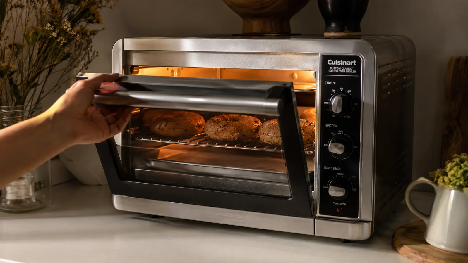A hand opening the toaster oven door to check golden bagels through the glass window