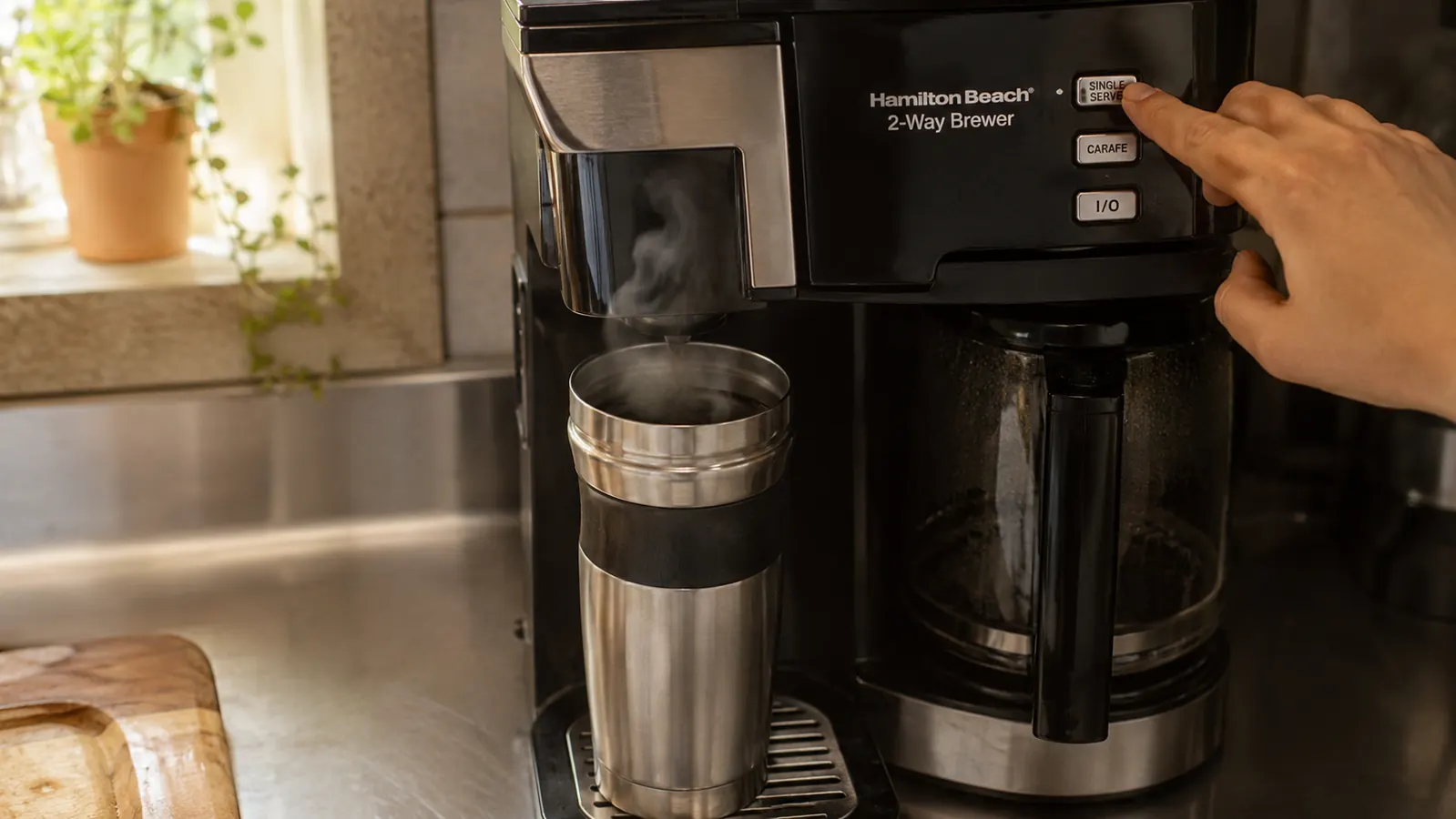 A hand pressing the single-serve brew button on the coffee maker while a travel mug sits underneath