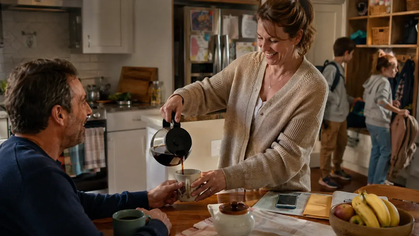 A mother pouring coffee for a husband seated at a small kitchen table while two kids grab backpacks nearby