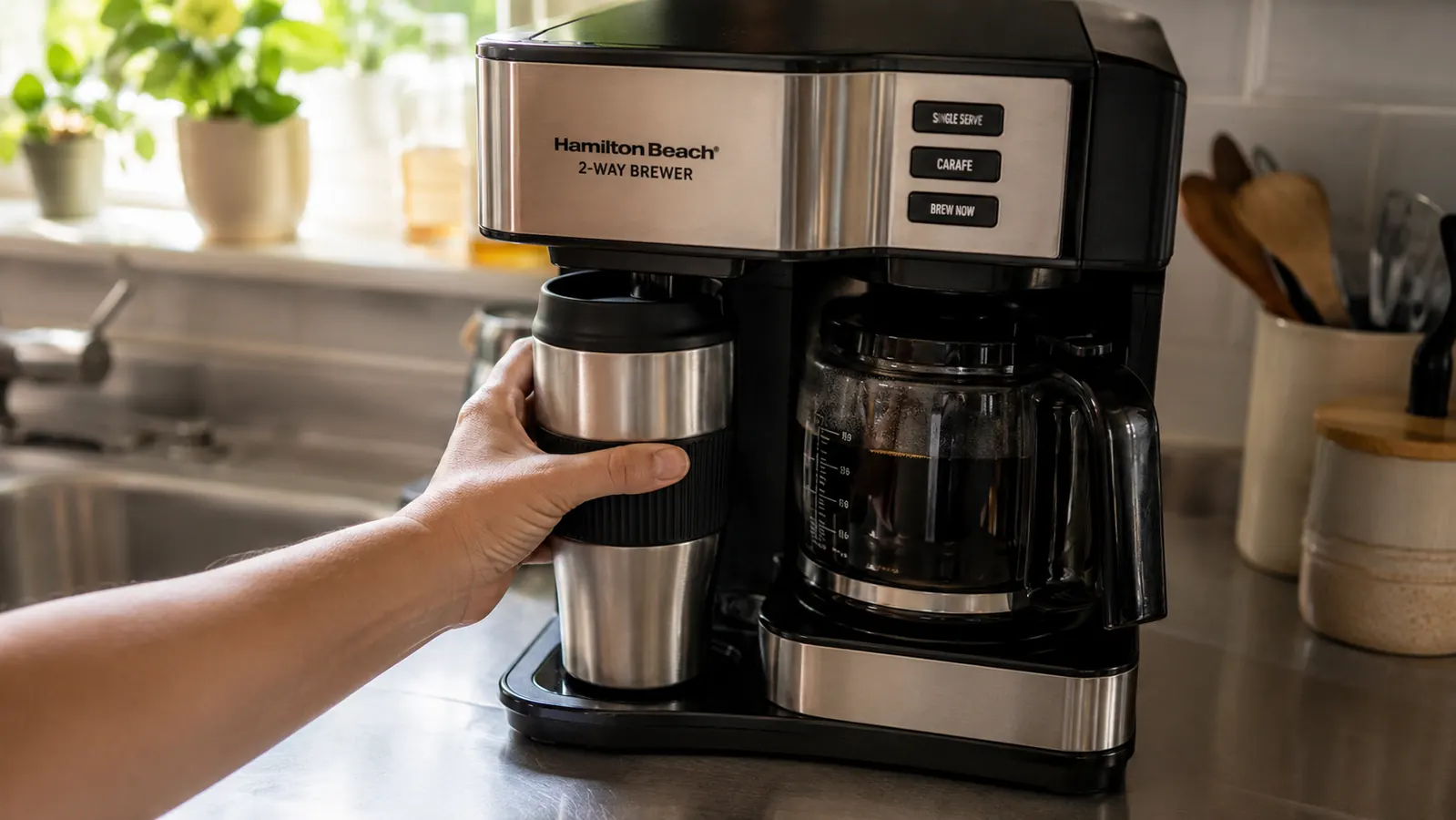 A hand placing a travel mug under the single-serve side of the 2-way brewer while a full carafe sits ready nearby