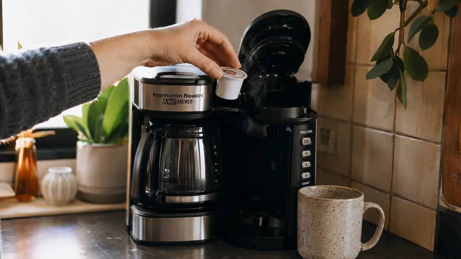 A hand loading a single-serve coffee pod into a two way brewer on a narrow kitchen counter