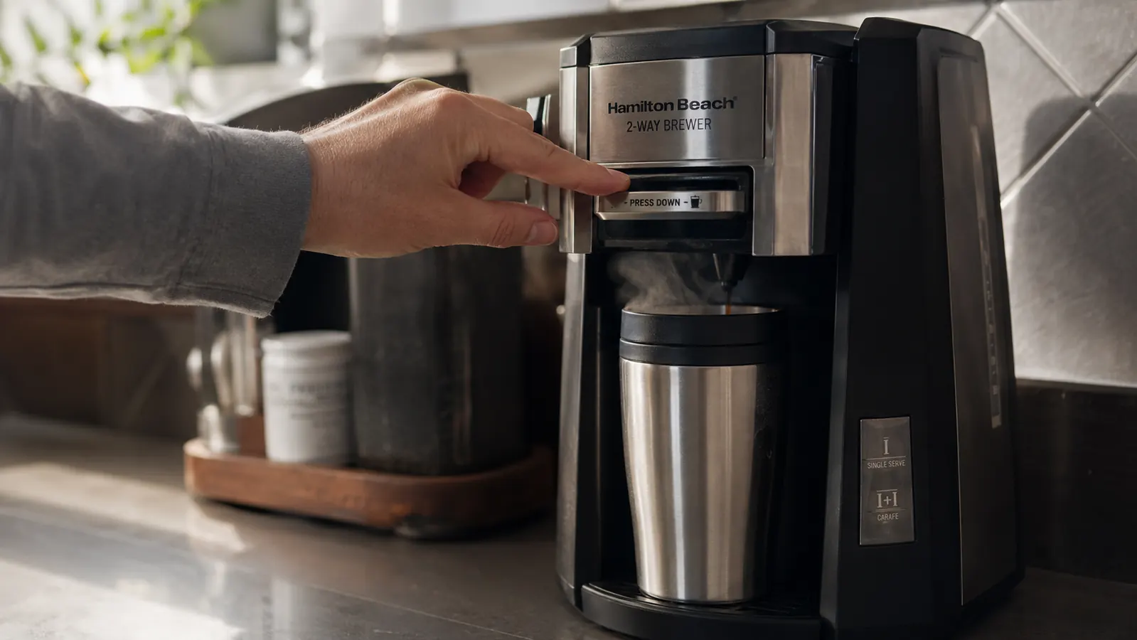 A hand pressing the single-serve brew lever on a coffee maker with a travel mug positioned underneath