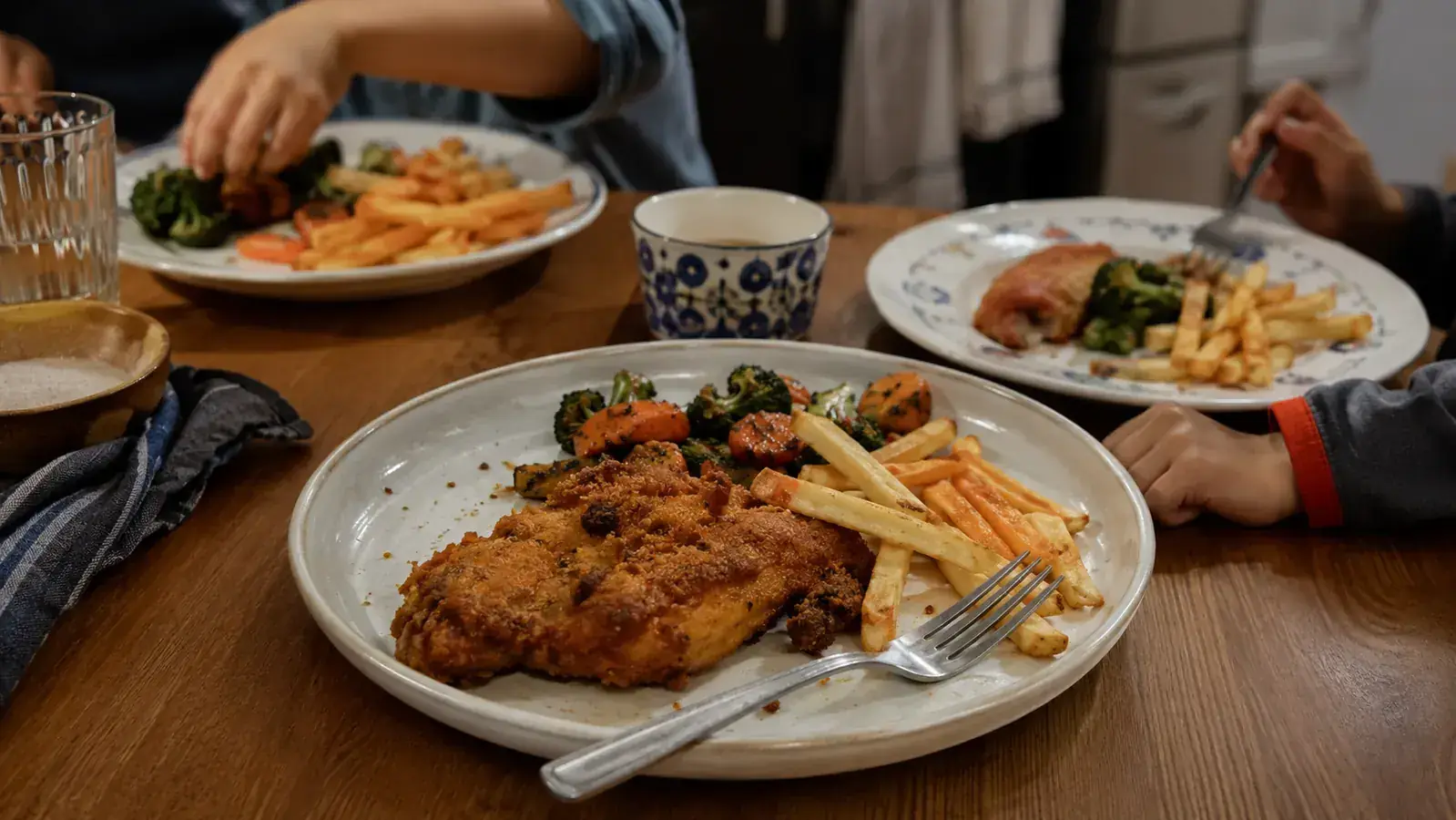 A family plate of crispy chicken, fries, and roasted vegetables served on a small kitchen table