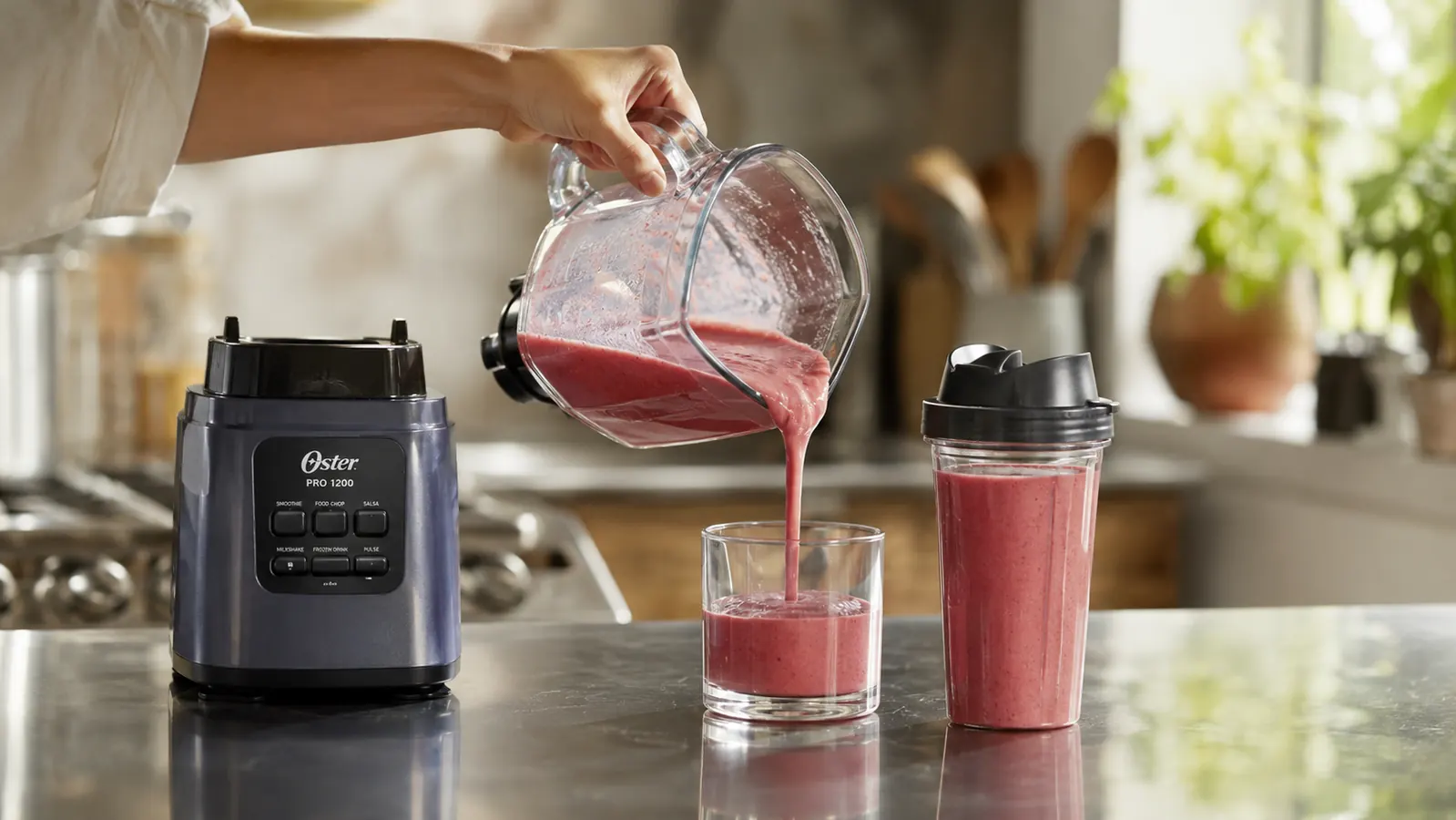 A hand pouring a full batch of smoothie from the blender jar into a glass and a to-go cup side by side