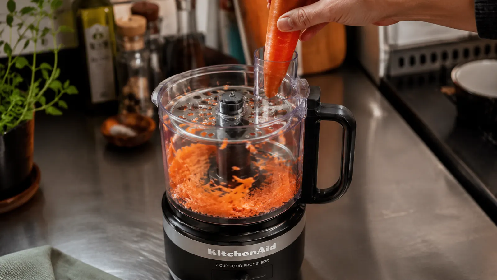 A hand feeding a carrot into the food processor's feed tube while the shredding disc runs