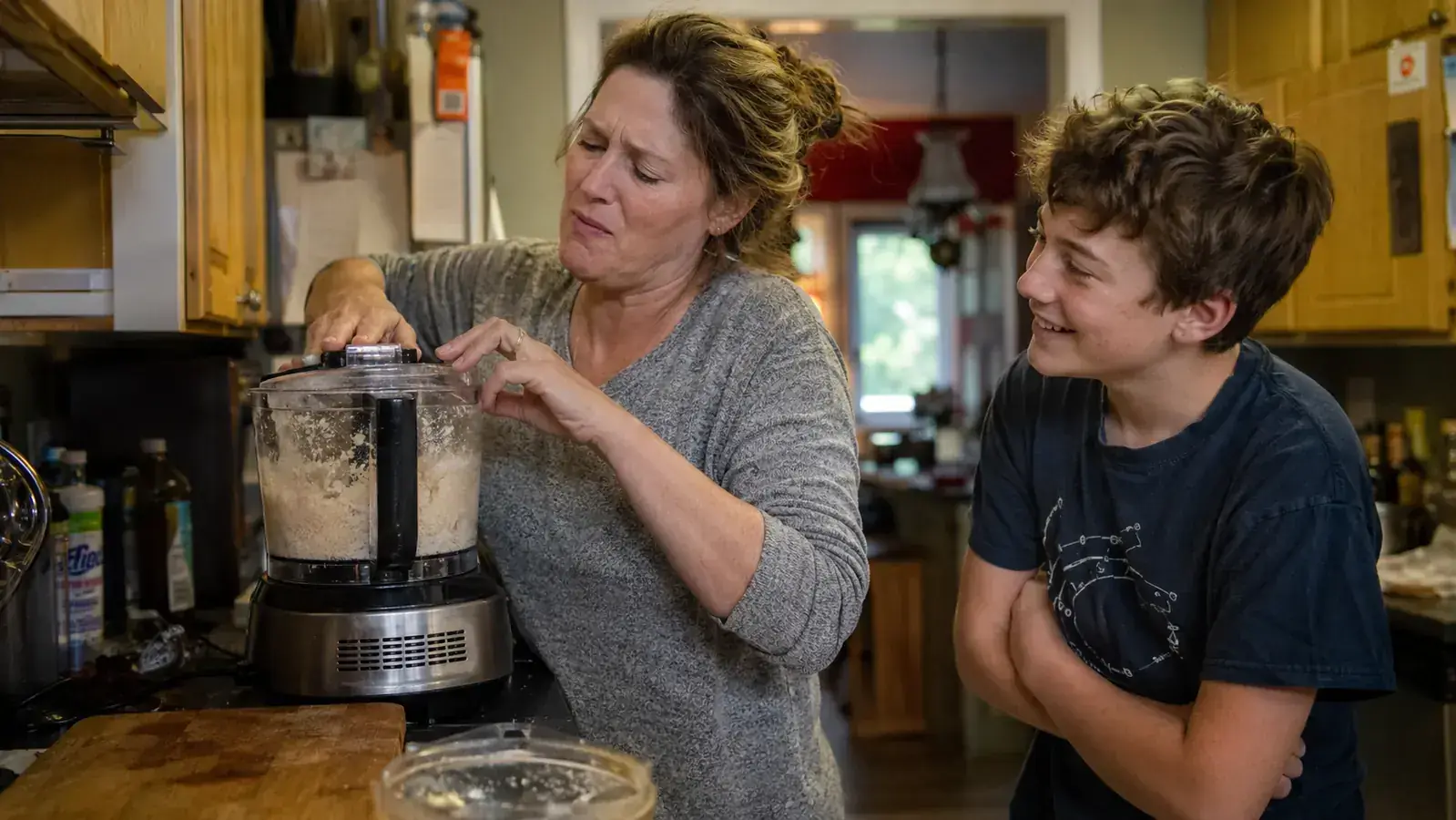 An 11 year old boy watching his mother wrestle with a food processor lid latch in a small kitchen