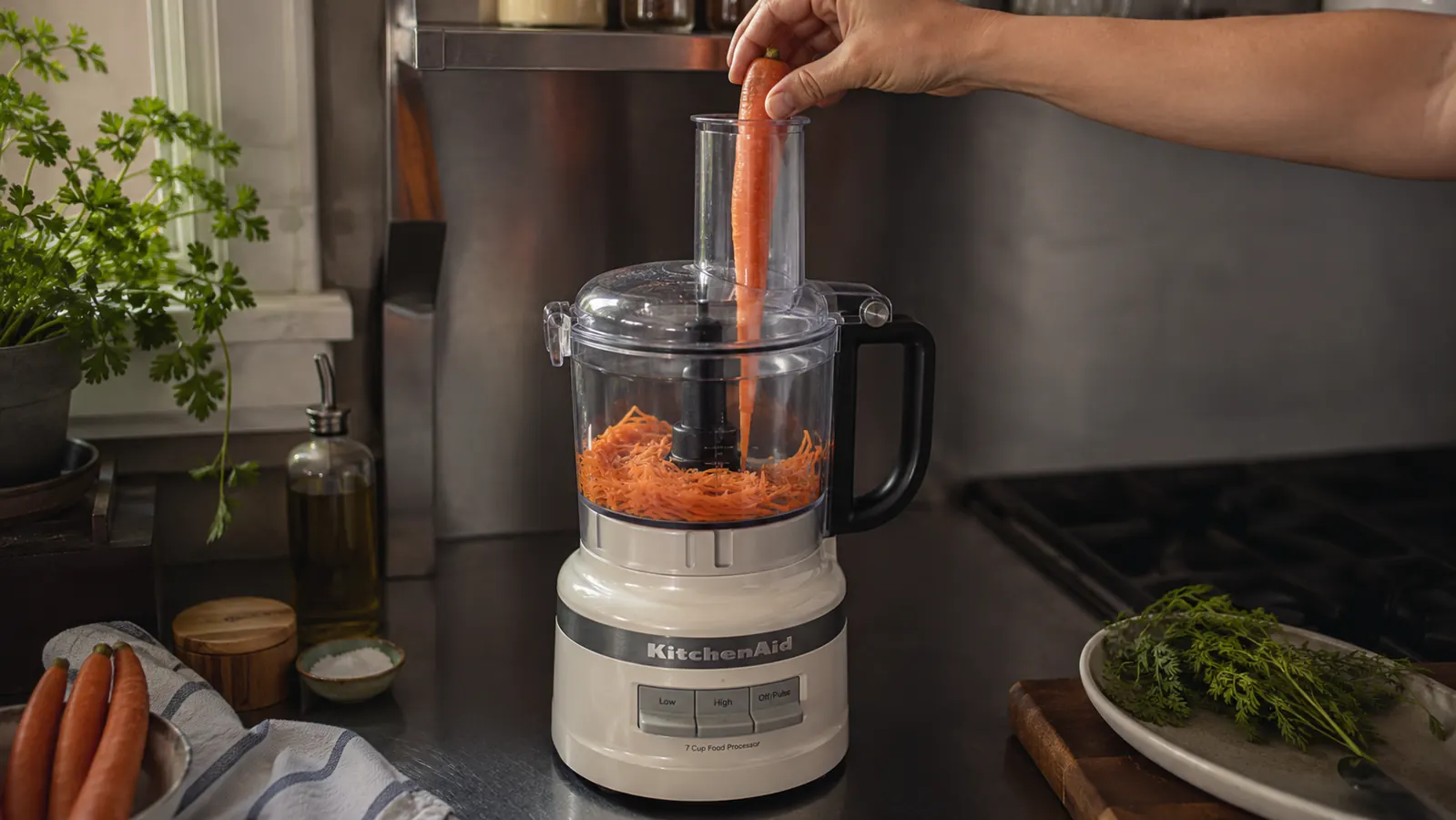 A hand feeding a whole carrot into the food processor's feed tube while the shredding disc runs below