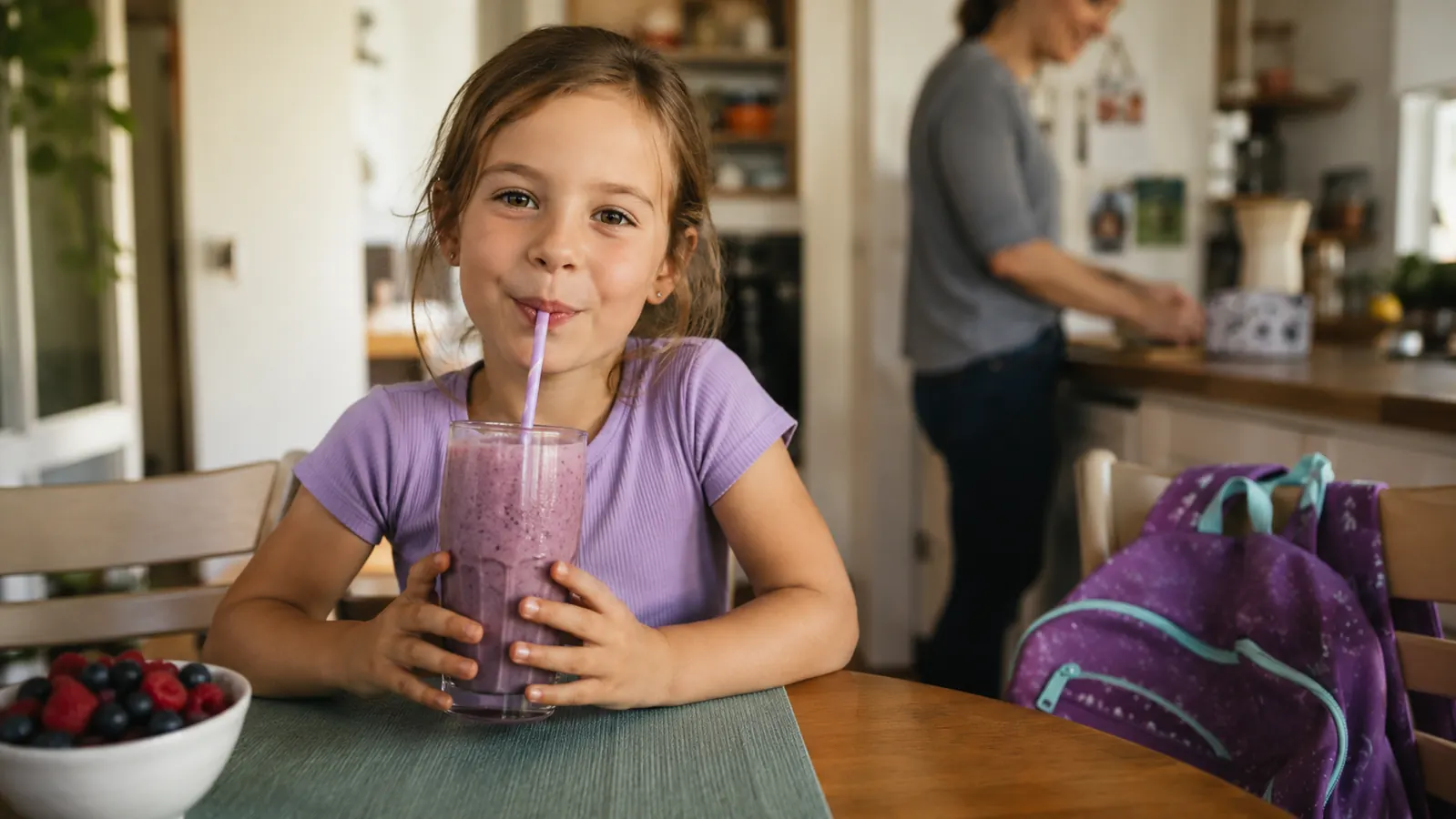 An eight year old girl drinking a purple smoothie at a small kitchen table before school